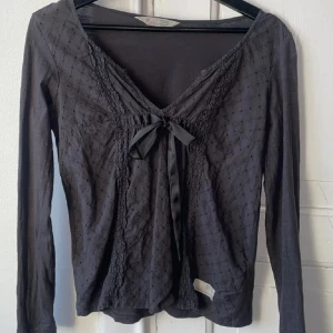 Odd Molly top - Supersöt grå långärmad blus med v-ringning och dekorativ rosett framtill. Blusen har broderade detaljer och spets längs urringningen, vilket ger en romantisk vibe. Tunn och mjuk i materiale.