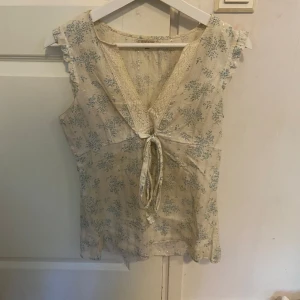 Blommig ärmlös blus med spets och knyt - Supersöt ljusbeige blus med små blå blommor, spetsdetaljer vid v-ringningen och knyt framtill. Ärmlös modell med volangkant på axlarna. Perfekt för vår och sommar när du vill ha en romantisk och bohemisk vibe.