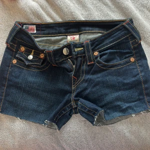 True Religion jeansshorts mörkblå - Snygga mörkblå jeansshorts från True Religion med ikoniska bakfickor, kontrastsömmar och råa kanter. Klassisk femficksmodell med dragkedja och knapp. Verkligen low waist på dessa