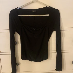 Svart långärmad topp från Glamorous - Svart långärmad topp från Gina tricot med fyrkantig urringning, liten rosett framtill och rynkade detaljer vid halsen. Toppen har croppad passform och vågig kant nertill och vid ärmslut. Perfekt för dig som gillar stilrena och trendiga plagg. Använd fåtal gånger och är som ny men har ingen användning för den. Storlek S🫶