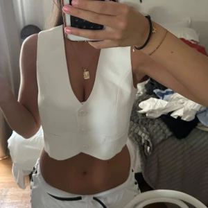 Vit croppad väst med knappar - Säljer en stilren vit väst med croppad passform och djup v-ringning. Västen har tre knappar framtill och en snygg, skräddad siluett. Perfekt att styla över en t-shirt eller på egen hand för en trendig look.