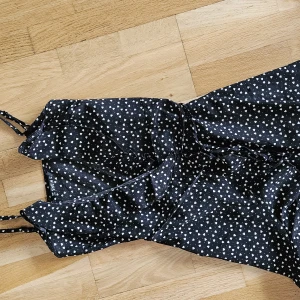 Svart omlott  hjärtprickig klänning m tunna axelband från SHEIN storl.S - Supersöt svart kortklänning från SHEIN med vita hjärtan över hela tyget. Klänningen har smala axelband, omlottkänsla och volangdetaljer nertill. Perfekt för dig som gillar lekfulla mönster och en feminin stil.