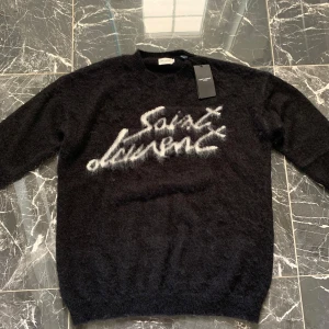 Svart Saint Laurent stickad tröja - Svart stickad tröja från Saint Laurent med vit logotyp i graffiti-stil framtill. Tröjan har lång ärm, rund halsringning och en fluffig, lyxig känsla. Perfekt för dig som vill ha en statement piece med exklusiv vibe.