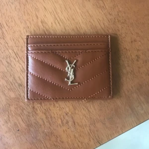 Brun kortfodral från YSL - Säljer ett snyggt brunt kortfodral från YSL i skinn. Det har en quiltad design och ett elegant YSL-emblem i guld på framsidan. Perfekt för att hålla dina kort organiserade med stil. Passar både till vardag och fest!