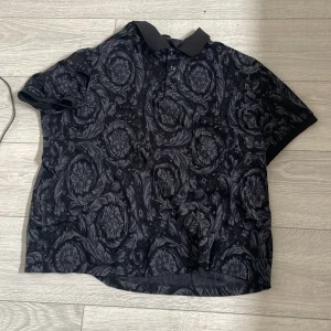 Svart pikétröja Louis Vuitton mönstrad - Exklusiv svart pikétröja från Louis Vuitton med grått, blommigt mönster över hela tröjan. Klassisk krage och knappar framtill. Perfekt för dig som vill sticka ut med en lyxig och unik stil.