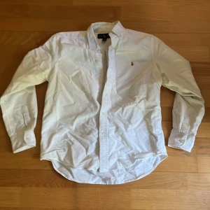 Vit skjorta från Ralph Lauren - Klassisk vit skjorta från Ralph Lauren med lång ärm och knappar framtill. Skjortan har den ikoniska broderade loggan på bröstet och är tillverkad i mjuk bomull. Perfekt för en clean och stilren look.