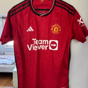 Manchester United fotbollströja  - Manchester Uniteds officiella hemmatröja från Adidas i rött med svarta detaljer och vita loggor. Tröjan har korta ärmar, svart-röda ränder på axlarna och klubbmärke på bröstet. Tillverkad i lätt och ventilerande polyester med diskret geometriskt mönster. Den är ifrån 2023/24 säsongen. 