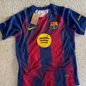 FC Barcelona Nike fotbollströja blå/röd - FC Barcelona matchtröja från Nike med klassiska blå och röda vertikala ränder, klubbmärke på bröstet och gul Nike-logga. Spotify-logga i gult på framsidan och UNHCR-tryck på ryggen. Tillverkad i Dri-FIT-material som andas. Kortärmad och sportig passform.