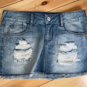 Blå jeanskjol med slitningar och glitter - Säljer en blå jeanskjol i kort modell med slitna detaljer framtill och rå kant nertill. Under de trasiga partierna finns glittriga inslag som ger kjolen en extra cool touch. Klassisk femficksdesign och bälteshällor. Perfekt för dig som gillar en edgy stil!