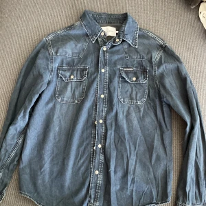 Jeansskjorta från Calvin Klein Jeans - Snygg jeansskjorta från Calvin Klein Jeans i klassisk blå denim. Skjortan har två bröstfickor med knappar, vit kontrastsöm och långa ärmar. Perfekt för en avslappnad och cool look. Passar dig som gillar tidlös stil och denim.