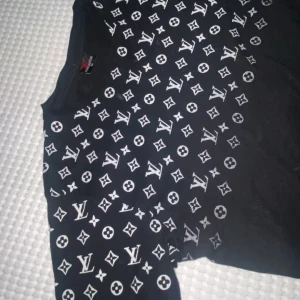 Svart Louis Vuitton t-shirt med logomönster - Svart t-shirt från Louis Vuitton i storlek M med ikoniskt vitt LV-logomönster över bröst och ärmar. Klassisk rund halsringning och korta ärmar. Materialet känns som bomull och passformen är normal. Perfekt för dig som gillar streetstyle och vill sticka ut.