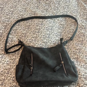 Svart axelväska i skinnimitation - Snygg svart axelväska med lång justerbar rem och flera dragkedjefickor både fram och bak. Väskan är rymlig med ett stort huvudfack och innerficka. Jag har köpt denna på Plick och vet ej vart den är ifrån därav priset. Jag har aldrig använt den och den såg i princip ny ut när jag fick den.