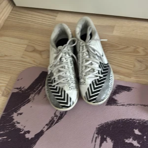 Nike vita och svarta fotbollsskor - Snygga fotbollsskor från Nike i vitt och svart med grafiskt mönster och stora swoosh-loggor på sidorna. Skorna har snörning och platt sula, perfekta för inomhusspel. Materialet är syntet och designen är riktigt modern och sportig.