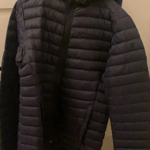 Mörkblå dunjacka Peak Performance 170 - Snygg mörkblå dunjacka från Peak Performance, modell JR Frost DH. Jackan har huva, dragkedja framtill och två fickor. Den är quiltad med horisontella sömmar och passar perfekt när det är kallt ute, perfekt till vintern och våren. Inga skador alls på jackan, ganska så ny