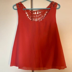 Röd ärmlös blus med spetsdetaljer - Snygg röd blus med ärmlös design och vackra virkade spetsdetaljer upptill både fram och bak. Luftig och flowy passform, perfekt för varma dagar. Blusen har rund halsringning och fina blommiga mönster i spetsen som ger en unik look.