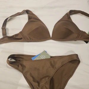 Ny brun bikini från H&M  - Snygg brun bikini från H&M med enkel och stilren design. Överdelen har vadderade kupor och justerbara axelband i stl 85 C. Underdelen är klassisk med låg midja i stl 42.Tillverkad i 80% polyester och 20% elastan för en skön passform. Perfekt för strandhäng eller poolparty.