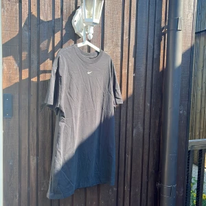 Svart Nike klänning i bomull - Svart klänning från Nike med klassisk vit swoosh broderad på bröstet. Enkel och stilren design med rund hals och korta ärmar. Tillverkad i mjuk bomull som känns skön mot huden. Perfekt för en chill och sportig look.