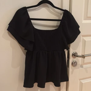 Svart blus med volangärmar - Supersöt svart blus med bred, smockad överdel och stora volangärmar. Toppen har en luftig, flowy passform och fyrkantig ringning. Perfekt för dig som gillar en romantisk stil