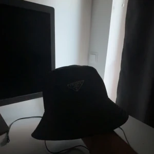 Svart bucket hat från Prada - Snygg svart bucket hat från Prada med klassisk trekantslogga framtill. Hatten är tillverkad i glansig syntet och har en stilren, enkel design som passar perfekt till streetwear. Insidan är fodrad för extra komfort och loggan finns även på etiketten inuti.