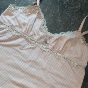 Ljusrosa randig linnetopp med spets - Supersöt ljusrosa och vit randig linnetopp med tunna axelband och spetsdetaljer längs urringningen. Dekorerad med en liten rosett och stjärnberlock framtill. Mjuk och skön, perfekt för sommarens varma dagar.