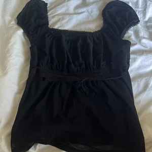 Svart blus med spetsdetaljer från SHEIN - Supersöt svart blus från SHEIN med korta puffärmar och spetskant runt halsringningen. Blusen har en markerad midja med knytband och fina spetsdetaljer framtill. Tillverkad i ett lätt och mjukt syntetmaterial som känns skönt mot huden.