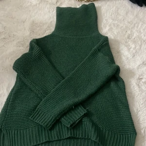 Mörkgrön stickad polotröja - Mysig mörkgrön stickad tröja med hög krage och långa ärmar. Tröjan har en chunky stickning och breda ribbade muddar vid ärmslut och nederkant. Perfekt för lager-på-lager och chill höst- och vinterstil.