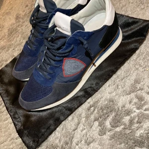 Philippe Model Trainers - Snygga blå sneakers från Philippe Model med detaljer i mocka och mesh, vit hälkappa och svart parti bak. Skorna har snörning och en ikonisk sköldlogga med röd kant på sidan. Perfekta för dig som gillar stilrena och sportiga vibes.