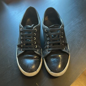 Svarta sneakers från Lanvin med lackad tå - Snygga svarta sneakers från Lanvin med klassisk rund tå och lackad tådel. Skorna har snörning och vit sula som ger en clean kontrast. Ovandelen är i mocka och detaljer i skinn, perfekt för dig som gillar stilrena och exklusiva sneakers.