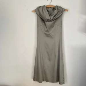 Silvrig  axelbandslös klänning - Elegant silvrig axelbandslös klänning med draperad krage och dragkedja bak. I mycket fint skick, har en mindre fläck på kragen (se sista bilden) som inte är märkbar och kanske går bort vid tvätt!