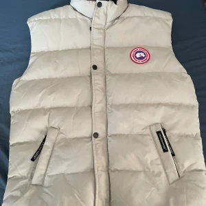 Beige dunväst från Canada Goose - Säljer en snygg beige dunväst från Canada Goose. Västen har en hög krage och stängs med knappar. Perfekt för kyliga dagar när du vill hålla värmen utan att kompromissa med stilen. Den ikoniska loggan syns både fram och bak. Storlek XL