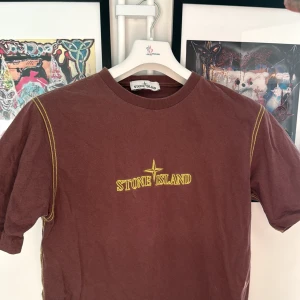 Brun t-shirt från Stone Island - Säljer en brun t-shirt från Stone Island med gul text på bröstet. T-shirten har korta ärmar och dekorativa sömmar i gult längs axlarna. 