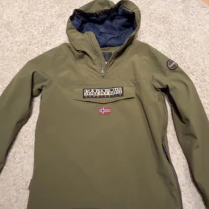 Grön jacka från Napapijri - Säljer en grön jacka från Napapijri med en praktisk huva och dragkedja. Jackan har en broderad logotyp och en norsk flagga på framsidan. Perfekt för utomhusaktiviteter med stil.