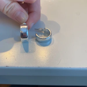 Silverfärgade örhängen - Säljer ett par stilrena silverfärgade örhängen i en klassisk rund design. Perfekta för att ge en elegant touch till din outfit. De är enkla att matcha med andra smycken och accessoarer.