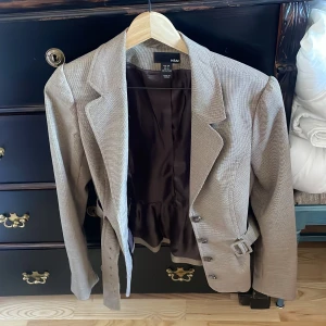 Grå kavaj från H&M - Snygg grå kavaj från H&M med ett diskret rutigt mönster. Kavajen har långa ärmar och ett bälte i midjan för en elegant look. Perfekt för en stilren outfit.