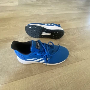 Blå Adidas sneakers - Snygga blå Adidas sneakers med vita detaljer och snörning. Perfekta för träning eller vardagsbruk. Skorna har en bekväm passform och en slitstark sula för bra grepp.
