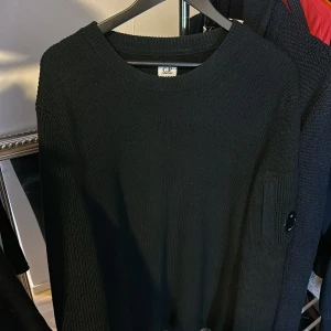 Svart stickad tröja från C.P. Company - Säljer en stilren svart stickad tröja från C.P. Company. Tröjan har långa ärmar och en rund halsringning. Den är dekorerad med en diskret logotypdetalj på ärmen. Perfekt för en avslappnad och trendig look.