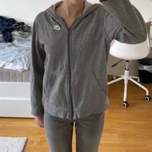 Grå huvtröja från Nike - Säljer en grå huvtröja från Nike med dragkedja och fickor framtill. Tröjan har en klassisk design med Nike-logga på bröstet och är tillverkad i ett mjukt material. Perfekt för en avslappnad stil.