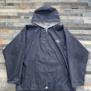 Marinblå/svart vindjacka från Calvin Klein - Säljer en marinblå/svart vindjacka från Calvin Klein med randigt foder och dragkedja. Jackan har en huva och två fickor framtill. Perfekt för blåsiga dagar. 💨