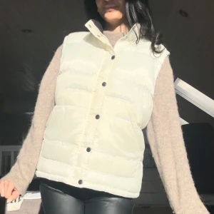 Vit dunväst med knappar - Snygg vit dunväst med svarta knappar framtill. Perfekt för lager-på-lager under kyliga dagar. Västen har en klassisk design med praktiska fickor. Dunet har dock samlat ihop sig lite klumpigt på vissa ställen, tydligen ska man kunna tvätta den med en tennisboll så ska det förmodligen lösa sig. 