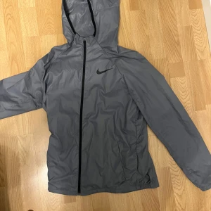 Grå vindjacka från Nike - Säljer en stilren grå vindjacka från Nike med huva och dragkedja. Jackan har en normal passform och är perfekt för blåsiga dagar. Den ikoniska Nike-loggan syns på bröstet. Passar bra till både jeans och träningskläder. Detta är barnstorlek XL (158-170)