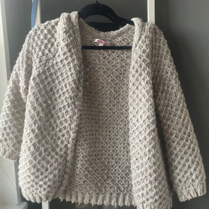 Beige stickad kofta från Indiska - Mysig beige stickad kofta från Indiska med grovt mönster och långa ärmar. Perfekt att slänga på sig över en topp för extra värme. Koftan har öppen front och ribbade muddar nedtill och vid ärmslut.