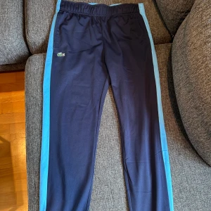 Mörkblå träningsbyxor från Lacoste - Snygga svarta träningsbyxor från Lacoste med ljusblå ränder längs sidorna och en vit detalj på baksidan. Byxorna har en elastisk midja för extra komfort och en liten Lacoste-logga på framsidan. Perfekta för en aktiv livsstil.