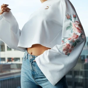 Vit croppad tröja från Puma * Kenza - Säljer en snygg vit croppad tröja från Puma med blommigt broderi på ärmarna. Tröjan har vida ärmar och ett guldfärgat Puma-logotyptryck på bröstet. Perfekt för en trendig look! 🌸