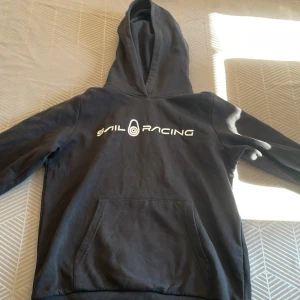 Svart hoodie från Sail Racing - Säljer en svart hoodie från Sail Racing med vit logga på bröstet. Perfekt för en avslappnad stil.