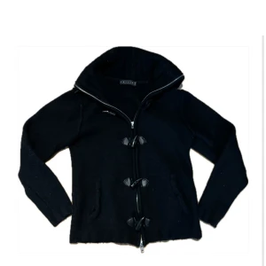 Duffel cardigan - Snygg svart stickad jacka med dragkedja och unika knappliknande detaljer framtill. Jackan har en hög krage och långa ärmar, perfekt för kyligare dagar. En stilren och mångsidig jacka för garderoben.