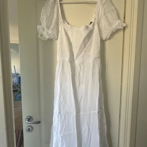 Vit långklänning med puffärmar - Charmig vit långklänning med puffiga kortärmar och smockdetaljer på överdelen. Klänningen har en söt knytning framtill och är perfekt för somriga dagar. 600kr nypris, aldrig använd.