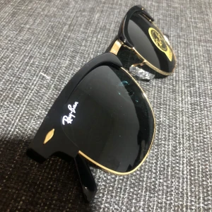Svarta och guldiga solglasögon från Ray-Ban - Snygga solglasögon från Ray-Ban med en klassisk design. Bågarna är svarta med guldiga detaljer som ger en lyxig känsla. Perfekta för att ge din stil en extra touch.
