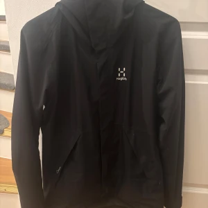 Svart vindjacka från Haglöfs - Säljer en svart vindjacka från Haglöfs med huva och dragkedja. Jackan har en stilren design och är perfekt för blåsiga dagar. Den är tillverkad i ett lätt syntetmaterial och har långa ärmar. Mitt pris är 1000kr. Vid frågor eller intresse, hör gärna av dig!