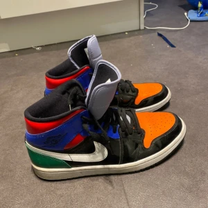 Flerfärgade Nike Air Jordans - Säljer ett par unika Nike Air Jordans i en mix av färger: orange, blå, röd, grön och svart. Skorna har en klassisk hög modell med snörning och vit sula. Perfekt för dig som vill sticka ut med en färgglad stil.
