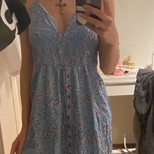 Blå blommig klänning - Söt blå klänning med blommigt mönster och röda knappar. Klänningen har en v-ringad design och knytband i nacken, vilket ger en öppen rygg. .